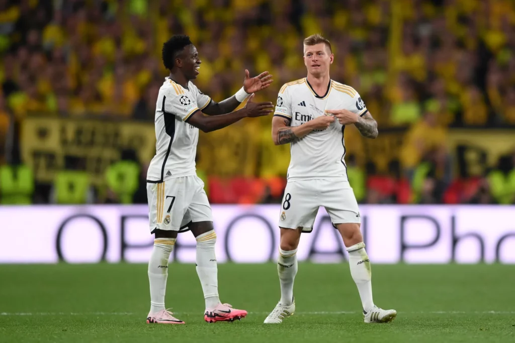 Toni Kroos, junto a Vinicius, se señala el escudo del Real Madrid.